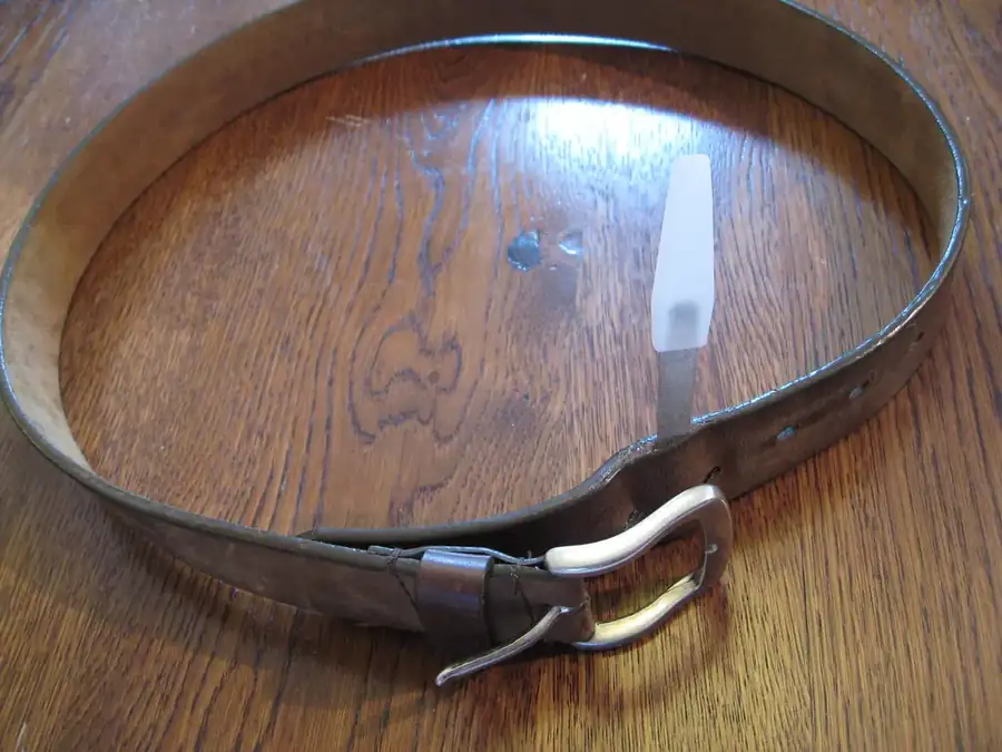 Cinturón de cuero oscurecido por el uso, apoyado sobre una mesa de madera, con la punta pasada por la hebilla